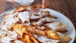 Dusting Angel Wing Cookies/ Polish Chrusciki with powdered sugar on a white platter