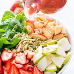 A fruit and spinach salad in a bowl with strawberry vinaigrette being poured on the salad