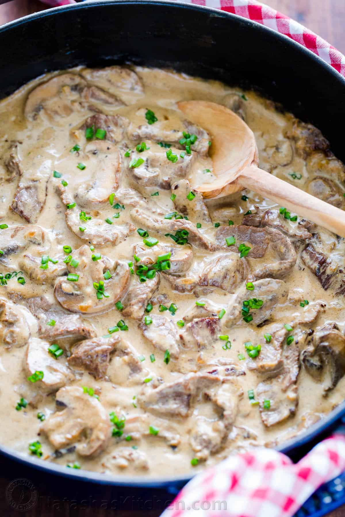 Classic Beef Stroganoff with tender strips of beef and mushrooms in an creamy Stroganoff sauce, served over noodles. An easy, one-pan, 30-minute recipe! #beefstroganoff #beefstroganoffrecipe #stroganoff #beef #beefrecipes #dinner #onepandinner #natashaskitchen Beef stroganoff with serving spoon garnished with chives