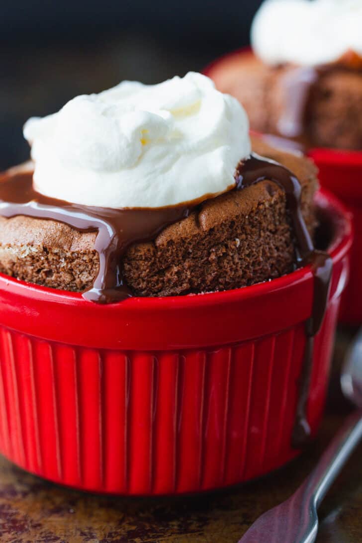 Chocolate Soufflé rises beautifully and is intensely chocolatey. Learn how to make Chocolate Soufflé with chocolate sauce and whipped cream. Red ramekin with chocolate souffle and toppings ready to serve