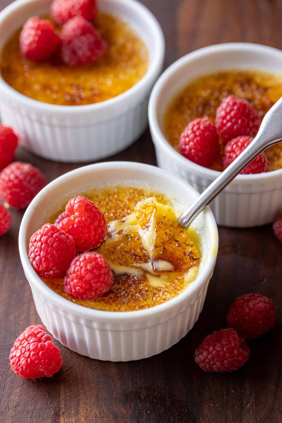 Creme Brulee is an easy dessert recipe made with just 4 ingredients! Learn how to make homemade creme brulee that tastes restaurant-quality. Three ramekins of creme brulee topped with raspberries, one with the caramelized sugar topping cracked with a spoon.