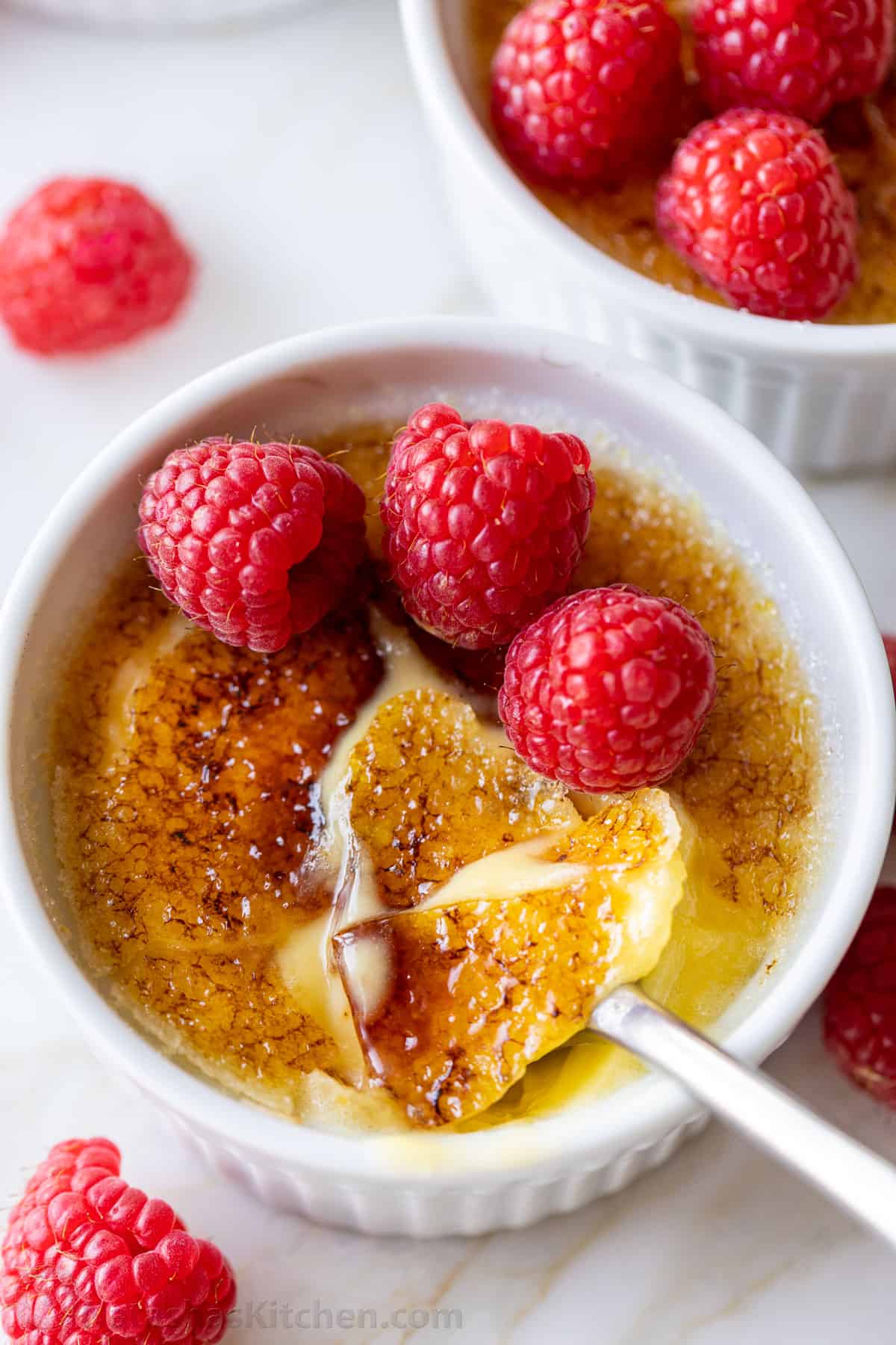 Creme Brulee is a creamy, indulgent dessert made with only 5 ingredients! Here's how to make a restaurant-quality vanilla creme brulee. Overhead view of creme brulee topped with raspberries, with the caramelized sugar topping cracked with a spoon.