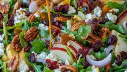 Pear Salad being dressed with Balsamic Vinaigrette in a white bowl, loaded with a variety of pears, red onion, goat cheese, cranberries, and toasted pecans