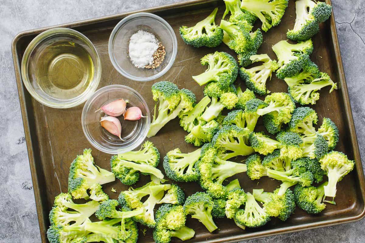 Ingredients needed for roasted broccoli are so simple; salt, pepper, oil and garlic. Raw broccoli florets with oil, garlic and seasonings in ramekins on a baking sheet.