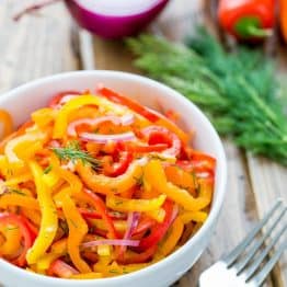 A bowl of zesty mini bell pepper salad with a fork, dill, mini peppers and purple onion beside it